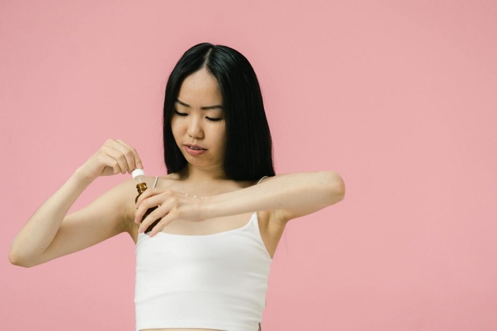 Asian woman using skincare product with pipette against a pink background, promoting self-care and wellness.