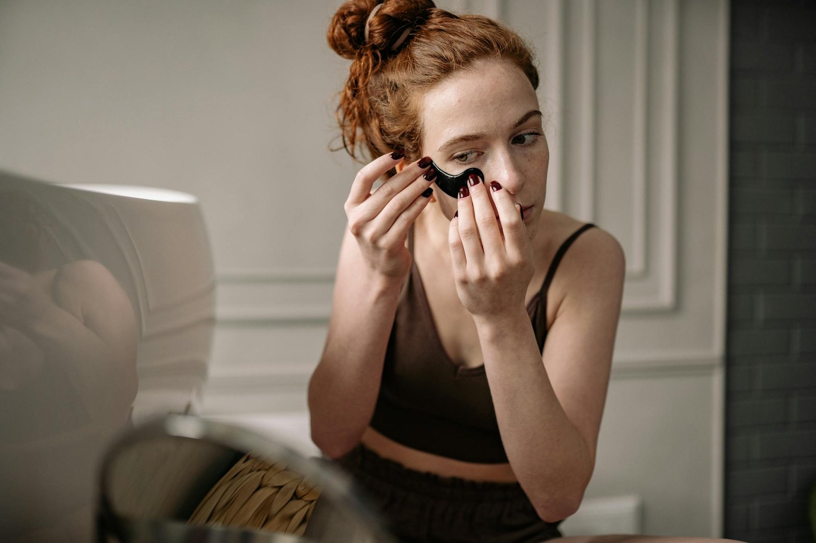 Red-haired woman applying an eye mask for skincare in a cozy room.