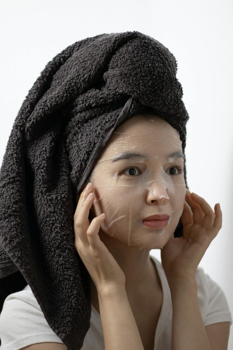 Woman with towel wrapped hair applying a skin care sheet mask indoors.