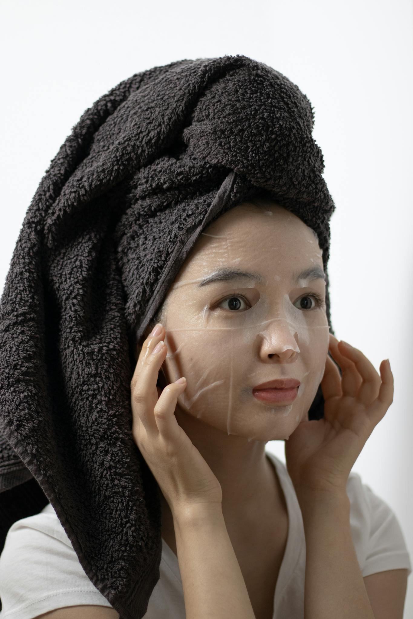 Woman with towel wrapped hair applying a skin care sheet mask indoors.